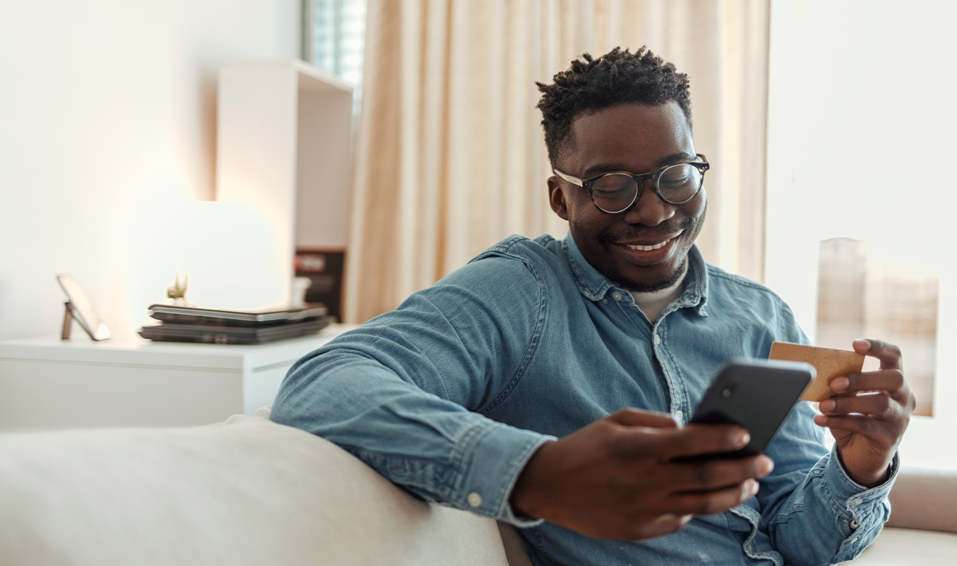 Man sitting on a sofa looking at a cell phone in one hand and holding a credit card in another.